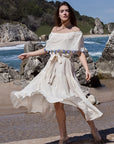 Front image of a woman wearing an off-the-shoulder cream beach dress, tied around the waist, 100% cotton with hand-knitted evil eye detail.