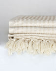 Close-up side image of a beige soft colour cotton blanket with a knotted fringe.