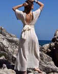 Back image of a woman wearing long cream 100% cotton summer maxi dress tied around the waist and with a low back.