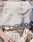 Close-up image of off-the-shoulder cream beach dress, tied around the waist, 100% cotton with hand-knitted evil eye detail.