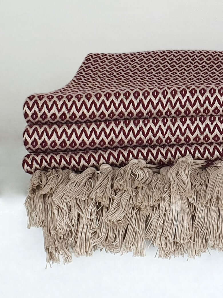 Close-up side image of a burgundy & beige colour cotton blanket with a tasselled edge.