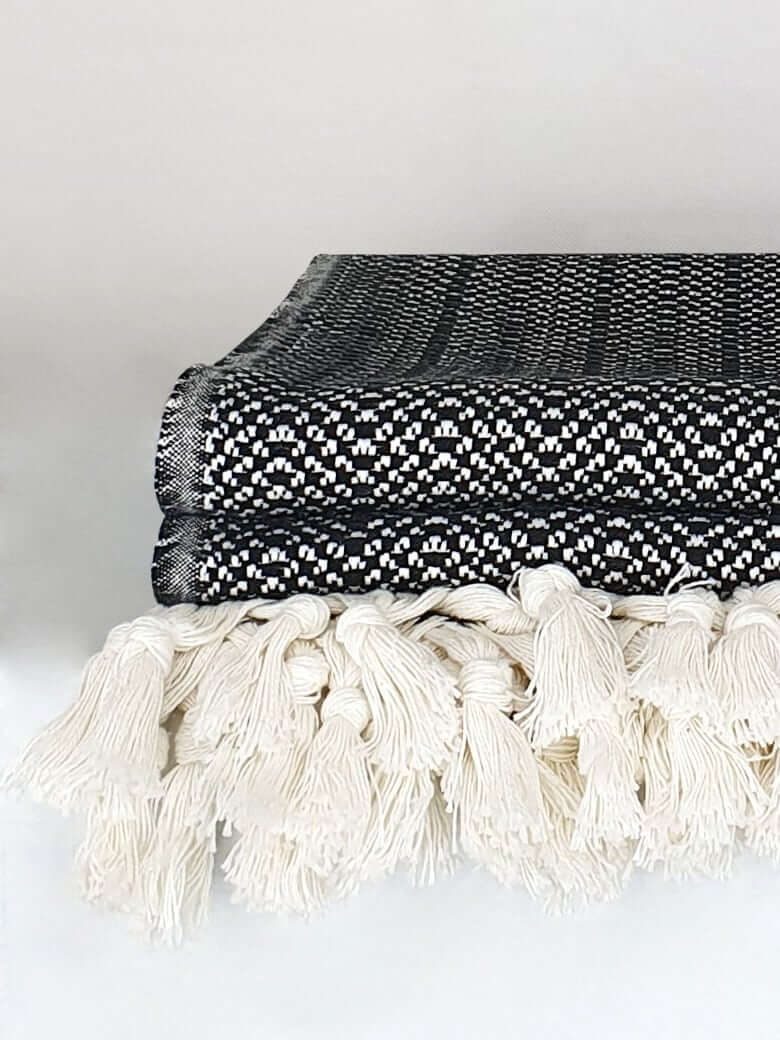 Close-up side image of a black & white colour king-size blanket with a tasselled edge.