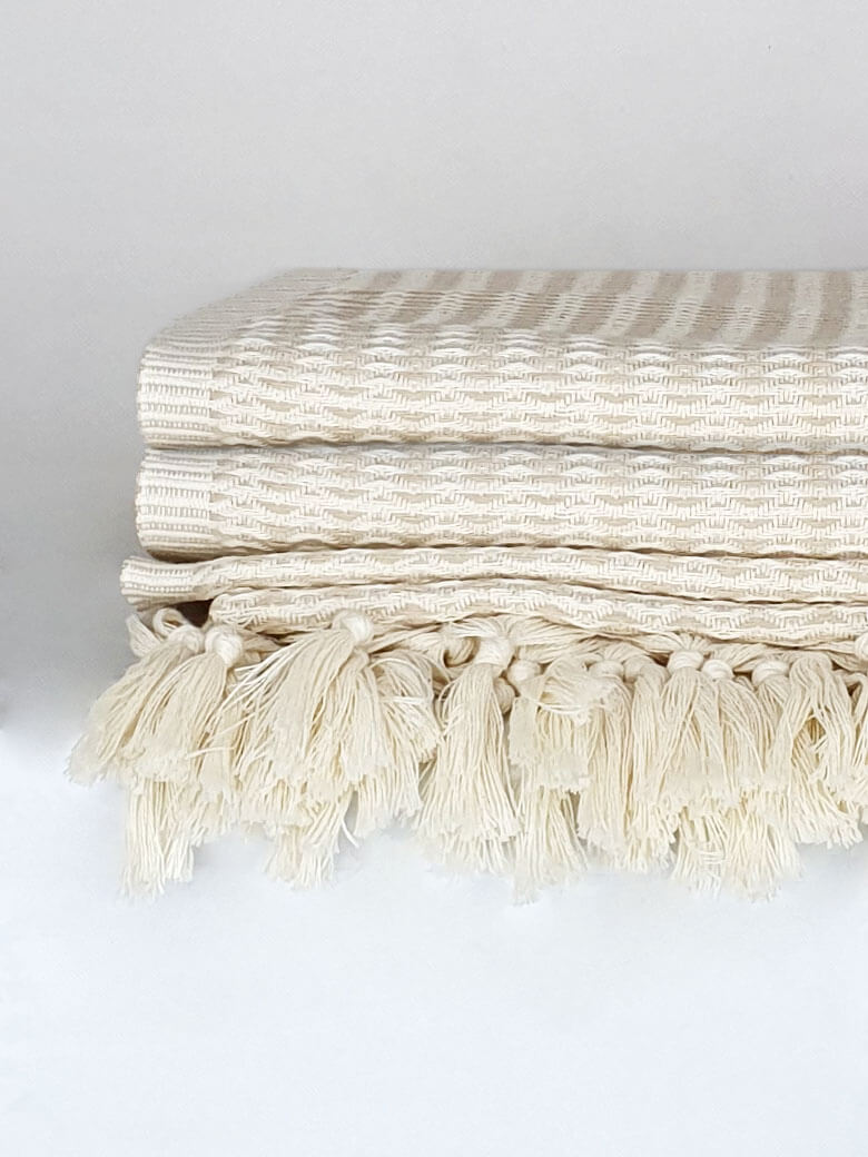 Close-up side image of a beige soft colour cotton blanket with a knotted fringe.