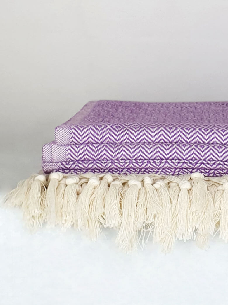 Close-up side image of a lilac & half part of a beige colour cotton blanket with a knotted fringe.