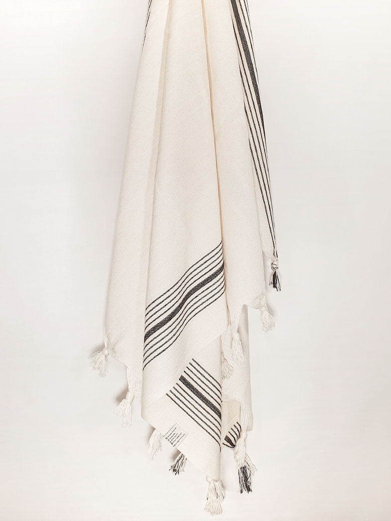 Plain beige hammam towel with vertical black stripes and knotted fringe hanging.