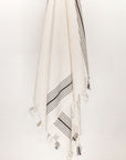 Plain beige hammam towel with vertical black stripes and knotted fringe hanging.